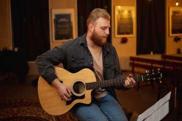 A young guy with a beard plays an acoustic guitar in a room with warm lighting