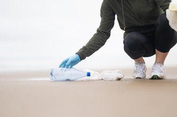 The hand of a female volunteer in blue gloves collects plastic bottles and garbage on the ocean shore. Environmental protection, poisoning of wildlife with poisons and toxins, recycling of garbage.
