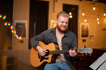Obraz premium A young guy with a beard plays an acoustic guitar in a room with warm lighting