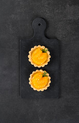 Twoo Tartlets with custard and slices of tangerines covered with syrup on a serving board on a dark gray background. Top view