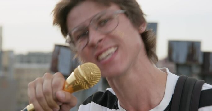 Young rocker guy with glasses is expressively singing with grimace using golden microphone on stand, his hair is disheveled by wind. Outdoor jam session. Blurred focus.