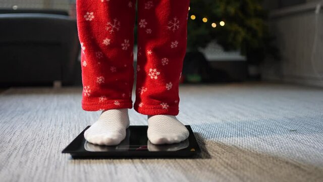 View of female feet climbing on electronic scale to control weight after Christmas holidays with Christmas tree in the background. Concept of bad nuricion at Christmas.