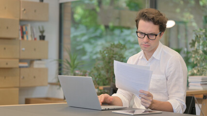 Man with Laptop Reading Documents in Modern Office