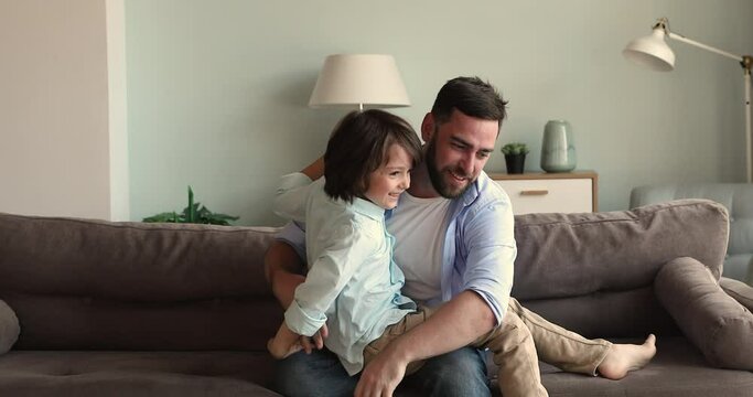 Affectionate laughing young dad elder brother sit on couch tickling tossing excited little boy son younger sibling. Active foster father small adopted child having fun messing giggling playing on sofa