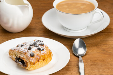 High angle view of homemade Danish Pastry filled with chocolate cream topping with chocolate chips and icing sugar in white ceramic dish and a white ceramic cup of milk coffee in white saucer.