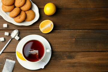 Flat lay composition with tea bag in ceramic cup of hot water, cookies and lemon on wooden table. Space for text