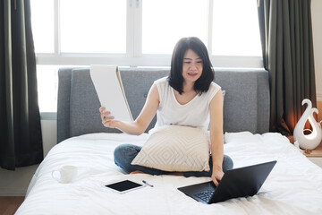 Asian young woman is sitting on white bed and reading papersheet for online working and learning...