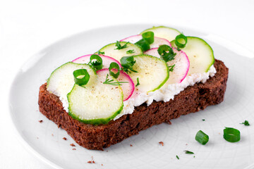 Sandwich with slices of radish and cucumber, cream cheese on the brown rye bread sprinkled with herbs on the ceramic plate. Vegetarian appetizer