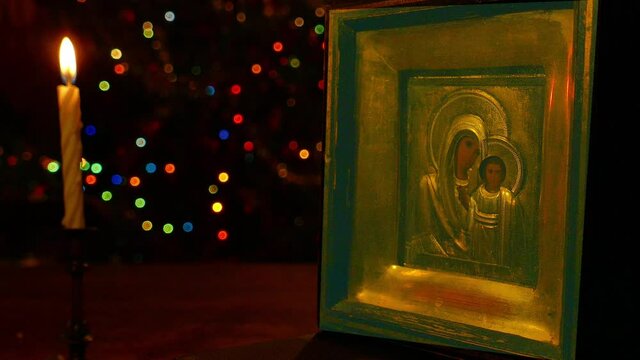 The icon of the Mother of God with the Christ Child is illuminated by a burning candle against the background of multicolored flickering garlanla lights