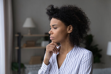 Lost in negative thoughts frowning stressed young african american woman touching chin looking in distance, considering personal troubles solution, suffering from psychological problems or loneliness.