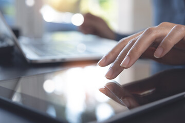 Close up of woman hand using digital tablet, finger touching on screen and working on laptop computer on table at home office