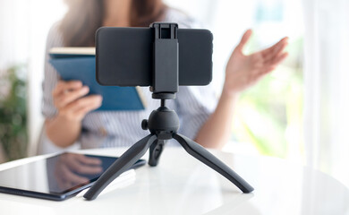 Focus on modern smartphone gadget on tripod, young asian woman in casual colthes  making live broadcast on Internet to coaching and working online at home, Concept working at home with new normal