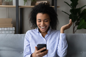 Fototapeta na wymiar Joyful laughing young african american woman looking at telephone screen, reading message with amazing news, feeling excited of getting online lottery betting gambling win notification at home.