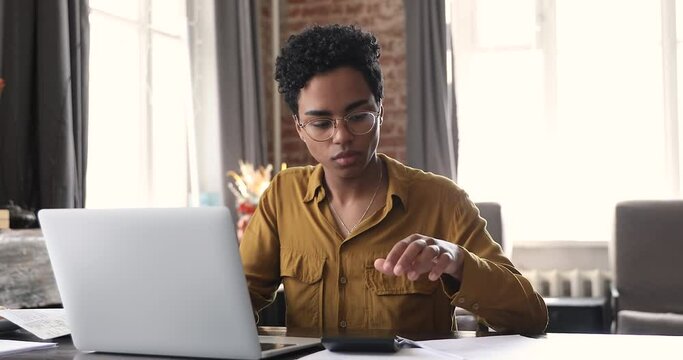 Serious African woman sit at desk do paperwork, reviewing household utility bills, calculates summary to pay, makes online payment through electronic bank app use laptop. Finances management concept