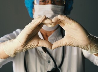 Kind elderly doctor in protective medical mask showing heart sign with hands in sterile gloves. The concept of a profession that saves lives and promotes charity.
