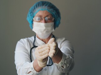 Photo of an elderly woman doctor wearing a mask in uniform putting on gloves against a gray background. Profession saving lives.