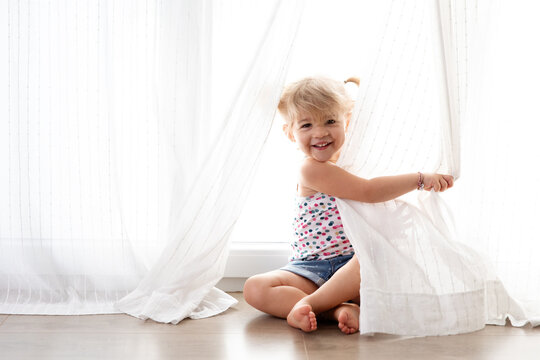 Happy toddler girl peeking behind curtains at home