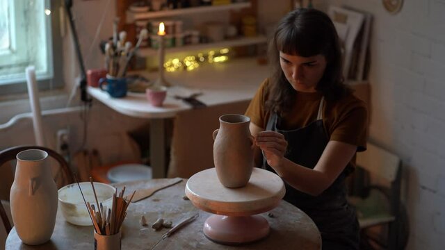 Woman creating pottery in cozy studio or workshop. Young female sit at table shaping clay jug. Professional ceramics artist at work. Girl producing potter for sale in craft kitchenware retail store