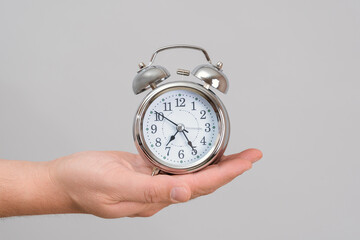 A man's hand holds a chrome alarm clock at arm's length, gray background