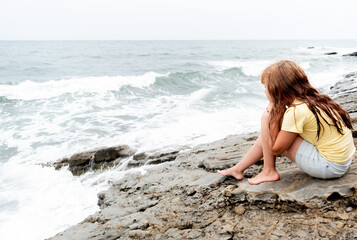 Fototapeta na wymiar Child girl sits on the rocks by the ocean. White waves. Adventure by the sea.