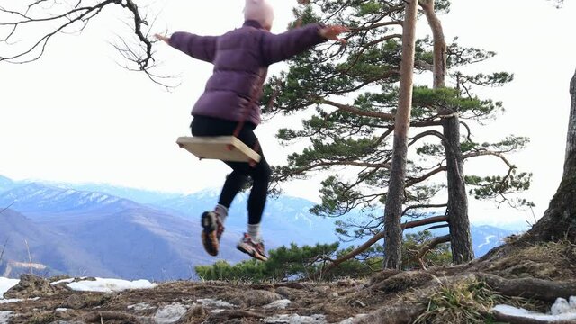 A girl in a purple jacket cheerfully rides a swing high in the mountains against the backdrop of a beautiful landscape of mountains and perennial trees, there is still snow.