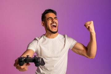 Fototapeta na wymiar Excited Arab guy winning online esports competition, playing video game, shouting OMG, making YES gesture in neon light