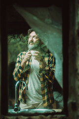 Mature adult man at home viewed from outside thrugh the glass winws drinking coffee and looking around - alternative rural lifestyle people hipster clothes and beard