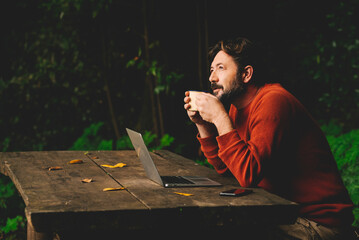 Concept of remote worker and smart working digital modern people lifestyle with adult hipster bearded man work outdoors on a table in the woods and drinking coffee thinking about job - online laptop