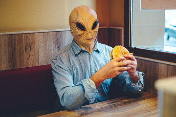 Extraterrestrial ufo alien eating an hamburger at the fast food sitting alone at the table -...