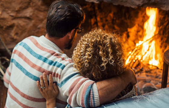 Romantic couple enjoy flame in front of fireplace in winter at home - cozy romance love man and woman viewed from back admiring fire to heat and enjoy relationship together
