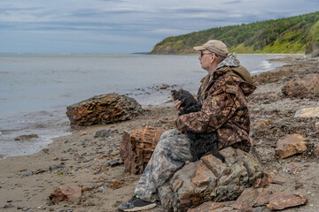 A pensive elderly man by the sea sits on a stone with a dog, thinking about the meaning of life.