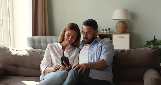 Friendly millennial spouses cuddle on sofa at home discuss online news at social media smiling on funny pictures internet memes on smartphone screen. Relaxed married couple enjoy web shopping on cell