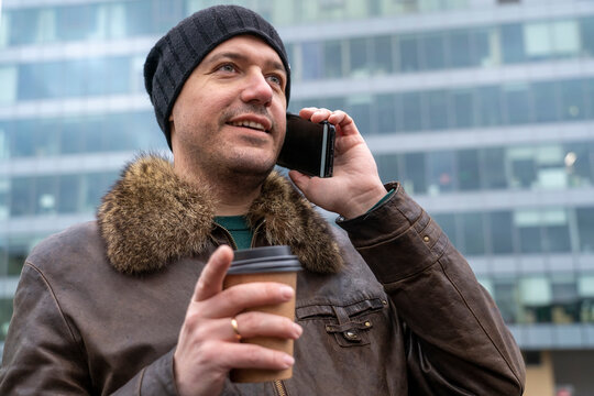 Millennial casual man wear black hat calling smartphone, stands outdoors at city street, holds paper cup of coffee togo, drinks takeaway hot tea. Guy enjoying coffee break while talking on mobilephone