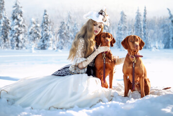 A blonde girl in a historical costume stands in the winter forest in the snow with two dogs. The...