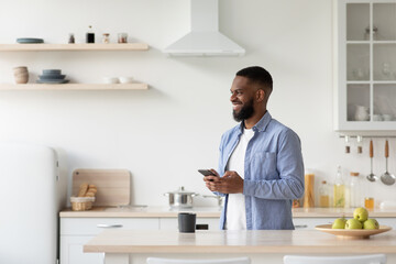 Glad young black man with beard enjoy good morning, typing on phone, looking at free space in modern kitchen