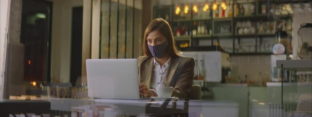 Adult Caucasian businesswoman sitting in restaurant or cafe and using laptop. Wearing textile face mask. Entrepreneur working outside office. - Powered by Adobe