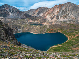 Fototapeta na wymiar Amazing view to beautiful blue mountain lake in bright sun. Heart shape blue alpine lake among sunlit green hills and large mountains under sun in cloudy sky.