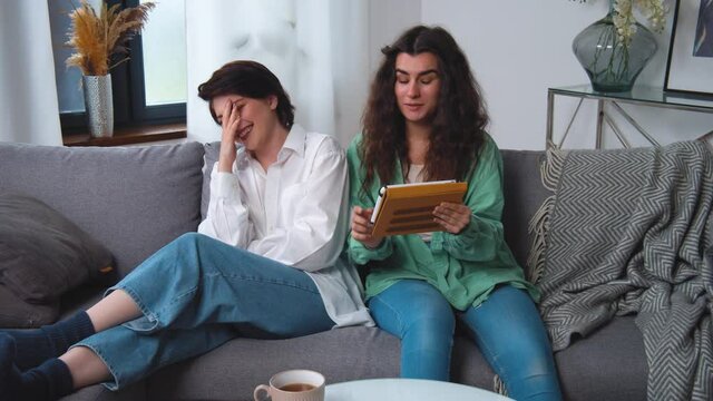Happy young lesbian couple couple sitting on sofa, using tablet computer, browsing and laughing at funny photos, videos, having fun together, enjoying their weekend at home. Caucasian women yelling