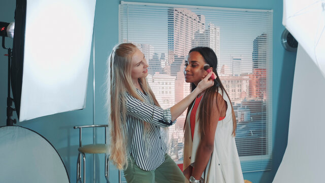 Behind the scenes on photo shoot: make-up artist preparing pretty black model to the photo shoot. There are skyscrapers in the background.