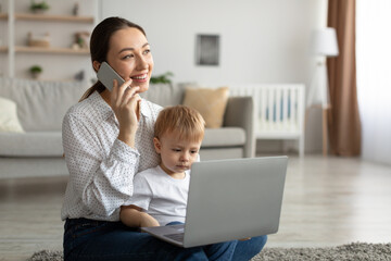 Happy mother with toddler son chatting on smartphone and using laptop computer, sitting on carpet...