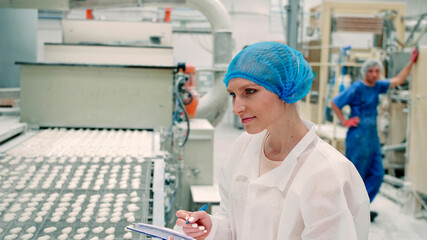 Candy factory. Controller checking conveyor with candies. Young woman in uniform holding folder and...