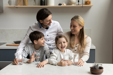 Happy parents and two healthy sibling kids drinking pure fresh clean water in kitchen, satisfying thirst, standing at table with glasses, smiling, laughing. Family care, children healthcare concept