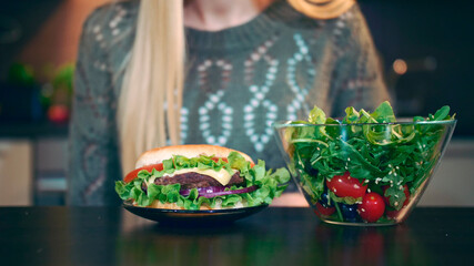 Crop view of Young lady preferring hamburger to salad. Attractive young woman choosing to eat healthy hamburger for breakfast while sitting at table in stylish kitchen.