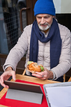 Confident grey haired man eating burger and typing at the tablet
