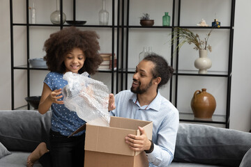 Happy Black dad and pre teen daughter kid unpacking parcel together, receiving package from...