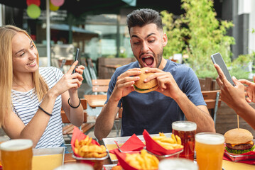 group of friends having fun at fast food restaurant, women use smartphone for shooting video and photos for social network, man eating junk food burgher