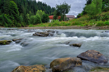 Long exposure of a mountain river with rocks from the bottom of the shot. With trees and a house in the background
