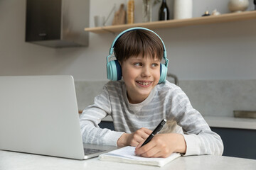 Happy gen Z teenage schoolboy with headphones and dental brackets writing learning notes at laptop, preparing essay, listening to audio course, webinar, looking at window away, thinking over home task