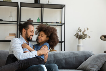 Happy cheerful Black dad cuddling, tickling playful excited daughter girl on couch, playing,...