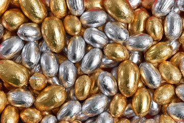 Large & small gold and silver foil wrapped chocolate easter eggs, against a pink, peach background.